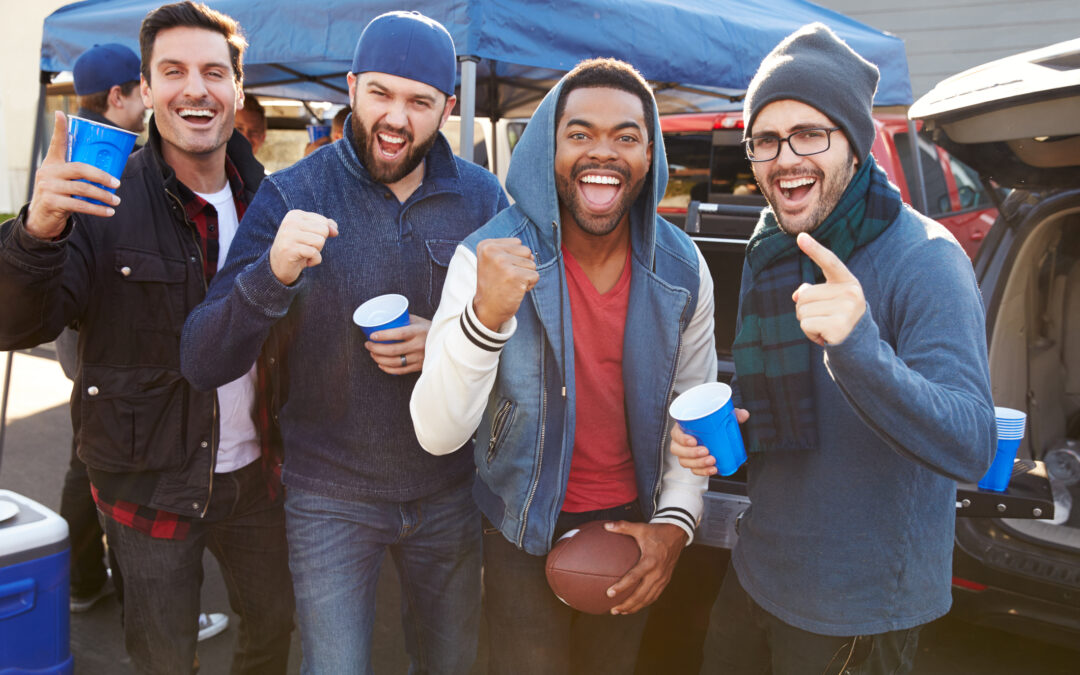 group of male sports fans tailgating in stadium car park with grill behind them