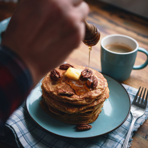 a hand putting syrup on a stack of pancakes. A coffee cup sitting in front of the plate and a fork to the right with a blue checked napkin beneath the teal plate.