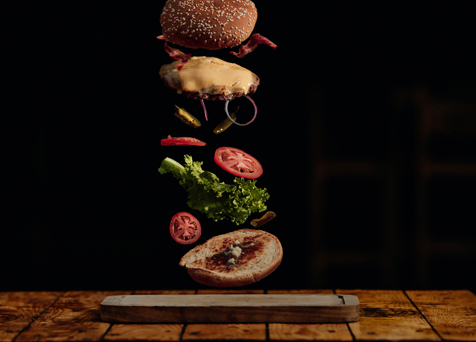 burger suspended in air with all the toppings and buns on a black background above a plate on a wood surface