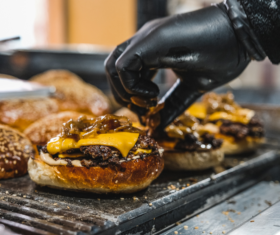 a gloved hand putting onions on smashburgers on the grill