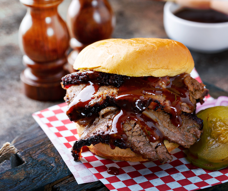 Shaved brisket on a bun with bbq sauce, sitting on a red and white checkered piece of paper with pickles on the side on top of a wood plank