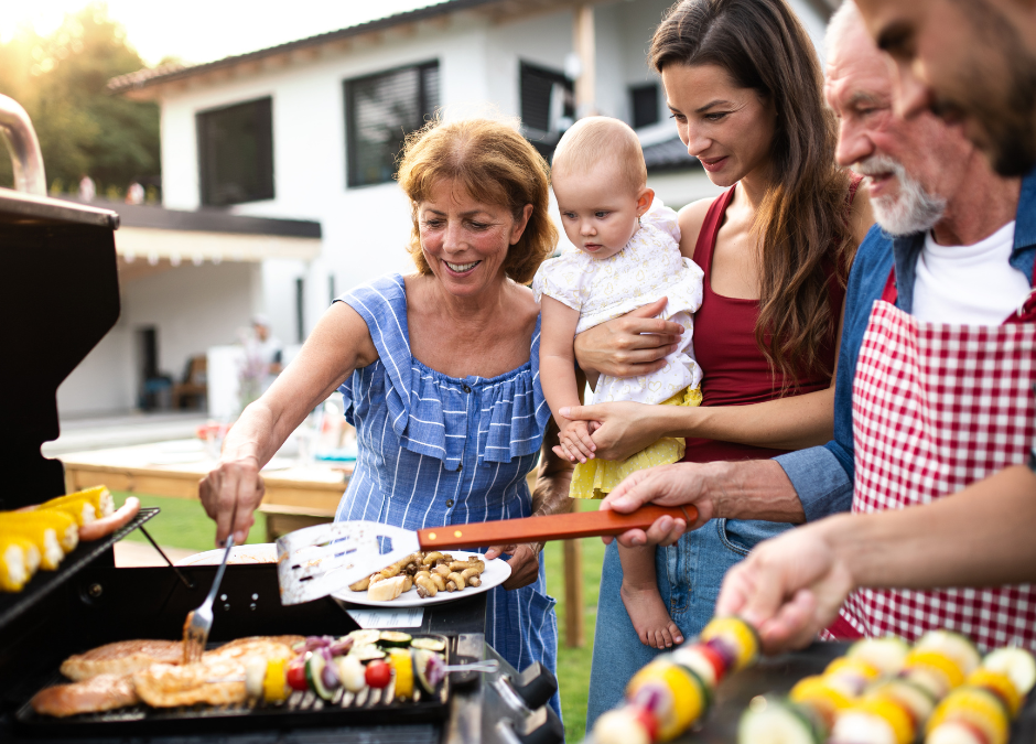 A family of 6 standing over a grill. A man with dark hair is putting kabobs on the grill, he is standing next to an older man, a woman holding a baby and an older woman