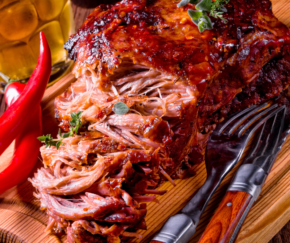 pork on a wooden board with half of it pulled. Two red peppers sit beside it with a fork and wood handled knife