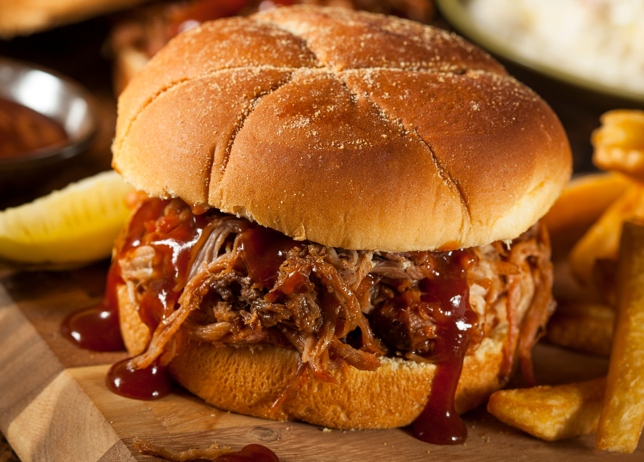 A pulled pork sandwich with bbq sauce dripping down the sides on a wooden board with fries piled behind