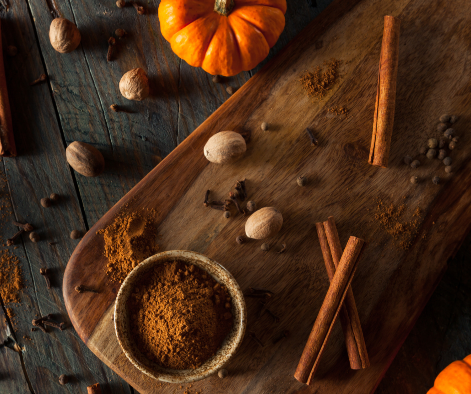 pumpkin spice in a small bowl on a wooden cutting board on a dark grey wood table with a small pumpkin, cinnamon sticks nutmeg and other spices