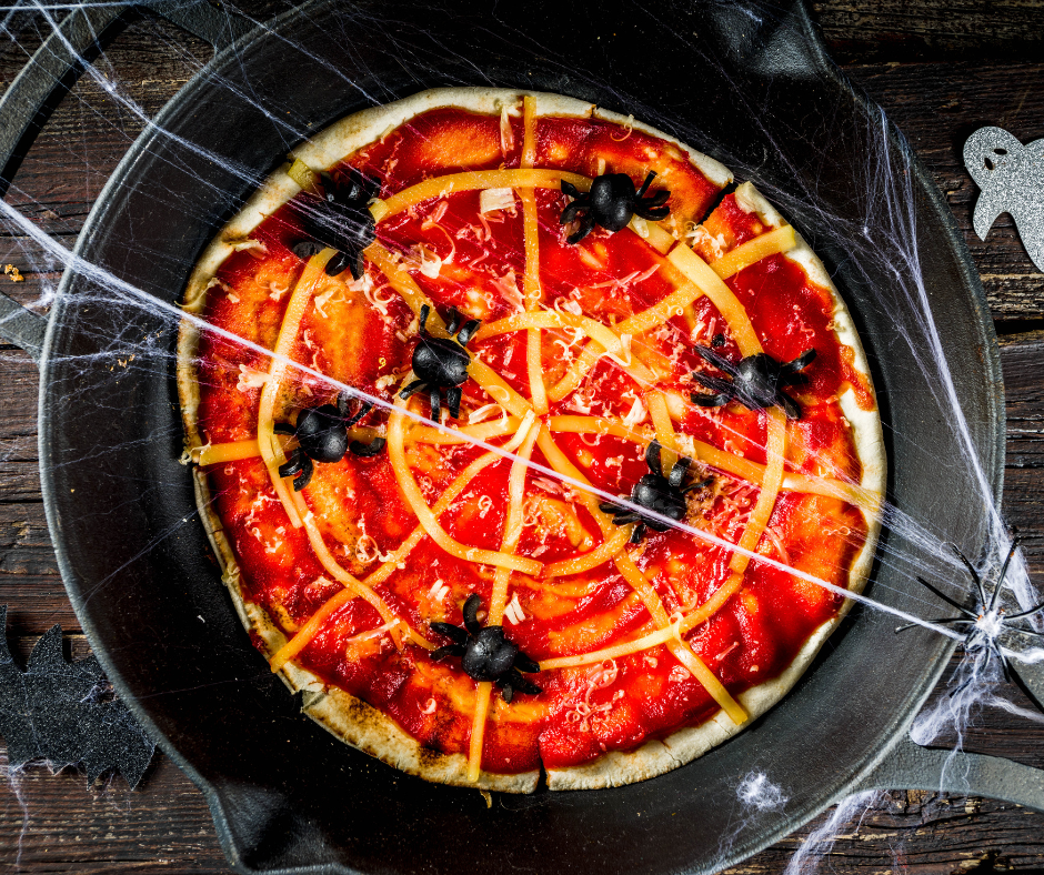 Spiderweb smoked pizza in a black cast iron skillet with olive spiders on a wooden table and a small ghost in the background